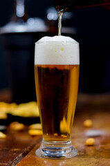 A glass full of beer in the foreground with a bottle of beer to the side and a black bucket with beers inside, some savory snacks on a wooden table with a black background.