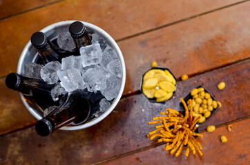 Black ice bucket with four beer bottles inside on a wooden table and with some savory snacks.