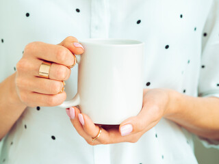 Mug mockup. Woman is holding white cup in hands. Wears white shirt and jewelry. Copy space for printing or branding