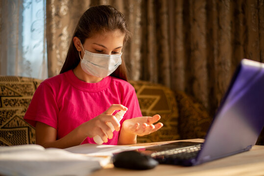 Small schoolgirl in medical mask working on the computer using hand sanitizer to keep hands clean