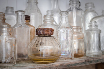 Collection of many dirty old glass bottles and flasks on shelf. All bottles are clear but have been exposed to the outside. Focus on one container with rusted lid / cap rusted and dirty brown liquid.