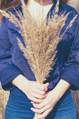 Toned vertical photo of young woman with long blond hair in blue shirt and jeans holding bunch of dried yellow bushgrass