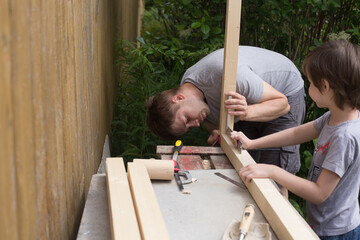 Obraz premium Boy assisting his father in DIY activities in the backyard of countryhouse. Man is working using chisel and boy is holding wooden billets. DIY and family time concept