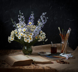 A bouquet of wildflowers stands on a wooden table, next to it is a mug of tea, a jar with artistic tassels, and an old book.