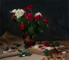 A bouquet of white and red roses stands in a clay jug on a wooden table. An old book is lying nearby.