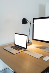 Devices on table with isolated screen for mockup. Computer blank display, laptop on home wooden desk. Lamps and white wall in background.