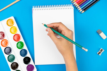 A schoolboy holds a pencil over a notepad on a blue background and supplies.