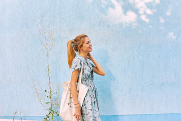 Young Caucasian woman in a blue dress with a bag near the blue wall in the summer street.