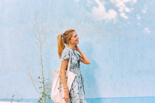 Young Caucasian woman in a blue dress with a bag near the blue wall in the summer street.
