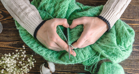 Warm cozy green sweater in the process of knitting and hands folded in the shape of a heart. Concept of your favorite hobby