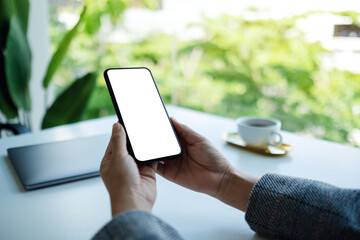 Mockup image of a business woman holding mobile phone with blank white desktop screen with laptop on the table