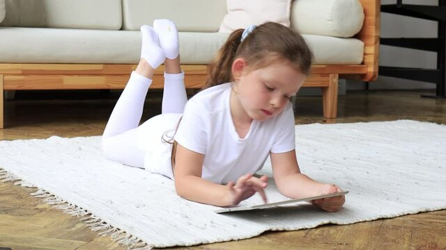 Child girl using digital tablet lying on carpet floor alone, holding and surfing touchpad internet at home.