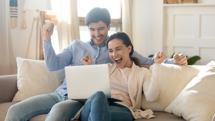 Overjoyed young married couple sitting on sofa, looking at computer screen, excited by good news in received unexpected email, feeling euphoric or celebrating online lottery win together at home.
