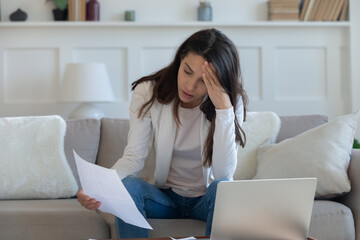 Stressed young lady holding banking paper termination letter, sitting on sofa indoors. Unhappy millennial woman reading eviction notification, thinking of financial problems, bankruptcy concept.