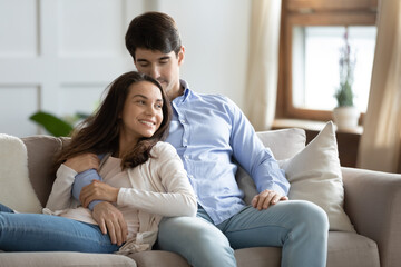 Obraz premium Affectionate loving young handsome man cuddling attractive smiling girlfriend, enjoying peaceful calm weekend time together on comfortable couch. Bonding happy couple relaxing on cozy sofa indoors.