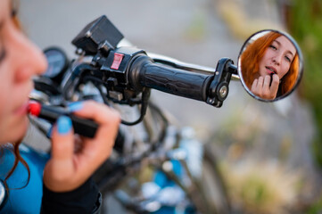 A woman on a motorcycle looks in the mirror and paints her lips with lipstick. Reflection of a red-haired biker girl.