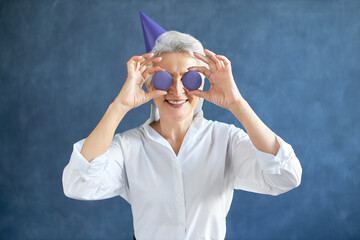 Horizontal image of happy gray haired retired Caucasian female in white shirt and conical hat holding two round almond cookies at her eyes, having fun. Playful mature woman posing with macaroons