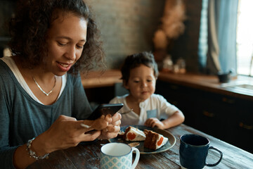 Busy young single mother using cell phone for distant work, writing posts on social network accounts, earning money from home on self isolation, little son sitting at table, eating cake in background