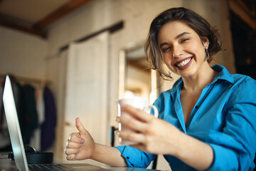 Portrait of cheerful beautiful student girl in blue dress smiling broadly, having fun while surfing internet on laptop, laughing at memes, holding cup of tea, making thumbs up gesture. Selective focus