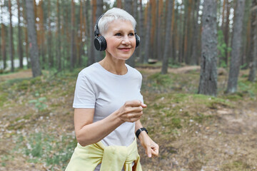 Active mature female runner in wearing sports clothes and wireless headphones, listening to music wearing headphones. Self determined energetic middle aged woman exercising outdoors, jogging