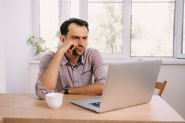 Angry man in shirt with a cup of coffee works on laptop