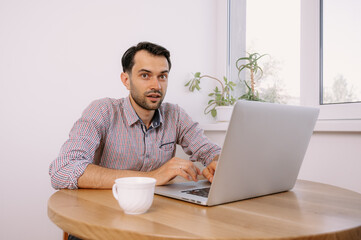 Happy smiling man in shirt with a cup of coffee works on laptop