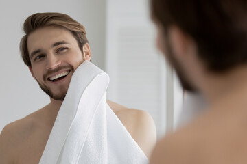 Mirror reflection smiling young man wiping face with white towel after shower or shaving, standing...