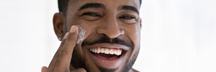 Close up head shot portrait overjoyed African American handsome young man applying moisturizing face cream, facial massage, enjoying skincare procedure, looking at camera, wide cropped image - obrazy, fototapety, plakaty