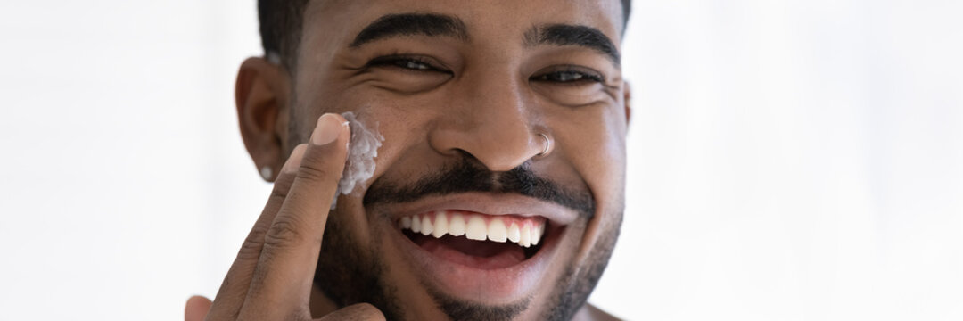 Close Up Head Shot Portrait Overjoyed African American Handsome Young Man Applying Moisturizing Face Cream, Facial Massage, Enjoying Skincare Procedure, Looking At Camera, Wide Cropped Image