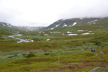 People on an hiking trail in a green valley in Sweden with a river, clouds and some snow