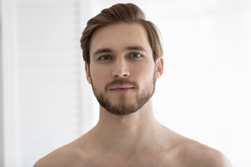 Obraz premium Head shot portrait handsome bearded young man with bare shoulders looking at camera, standing in bathroom at home after shower, personal hygiene, morning routine and skincare concept