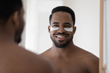 Close up head shot smiling African American young man with moisturizing hydrogel eye patches looking in mirror, standing in bathroom at home, enjoying skincare procedure, morning routine