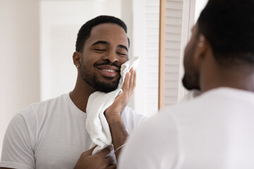 Mirror reflection close up satisfied smiling African American handsome young man wearing white t-shirt wiping face with soft towel after shower or shaving, enjoying morning routine in bathroom