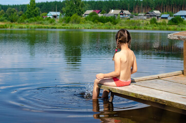 Baby girl sitting on a small bridge and splashing her feet in the river. A six-year-old child on summer vacation in the village.