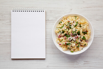 Homemade Macaroni Salad in a white bowl, blank notepad on a white wooden background, top view. Flat lay, overhead, from above. Copy space.