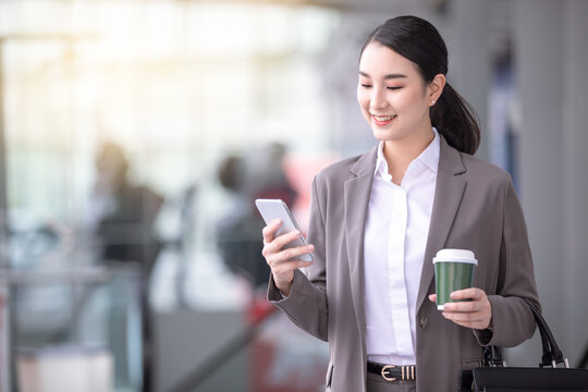 Asian woman with smartphone standing against street blurred building background. Fashion business photo of beautiful girl in casual suite with phone and cup of coffee