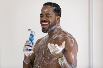 Smiling cheerful African American young man singing in shower, holding plastic bottle of...