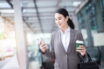 Asian woman with smartphone standing against street blurred building background. Fashion business photo of beautiful girl in casual suite with phone and cup of coffee
