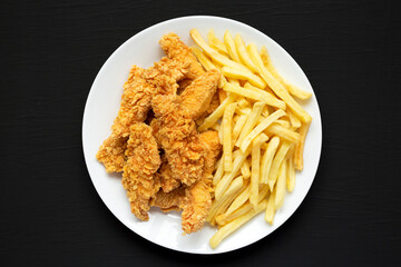 Homemade Crispy Chicken Tenders and French Fries on a white plate on a black background, top view. Copy space.