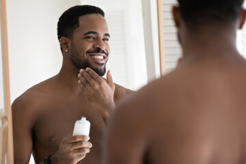 Mirror reflection close up smiling satisfied African American young man applying aftershave moisturizing lotion, standing in bathroom, enjoying skincare routine procedure after shaving - obrazy, fototapety, plakaty