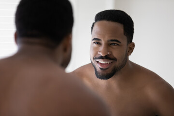 Fototapeta na wymiar Close up head shot smiling satisfied bearded African American young man with naked shoulders looking in mirror reflection, standing in bathroom, enjoying morning routine, skincare procedure