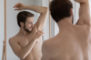 Mirror reflection young man applying antiperspirant on armpit after shower, standing in bathroom,...