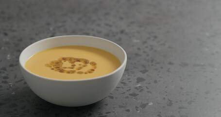 pour olive oil with spices to pumpkin cream soup in white bowl on terrazzo countertop