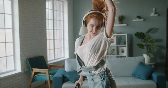 Funny crazy girl dancing with a vacuum cleaner. Joyful cute caucasian woman listening to music while cleaning her house - real happy people 4k footage