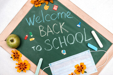 Blackboard with the inscription WELCOME BACK TO SCHOOL,flowers and protective face mask.Back to school concept in a new coronavirus reality.New normal concept.Flat lay,top view.