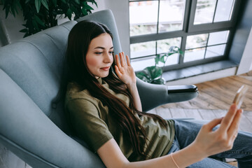 Attractive happy young woman relaxing in the home, sitting in the chair in living room, using smartphone, speaking on video chat, waving