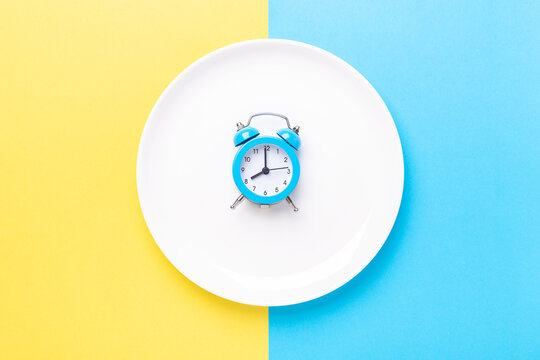 Blue alarm clock and empty plate on colored paper background. Intermittent fasting concept