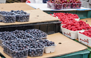 Small plastic boxes with blueberries displayed on food market, closeup detail, more raspberries and cherries background