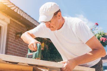 Carpenter old man using drill making holes in a wooden plank or electric saw cutting wood. Woodworking, home improvement, do it yourself (DIY) and construction works concept, action sunny shot.
