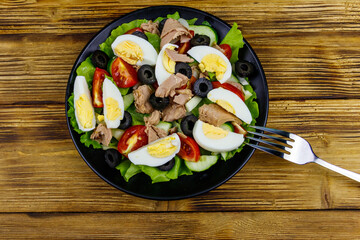 Tasty tuna salad with lettuce, black olives, eggs and fresh vegetables on wooden table. Top view